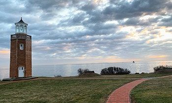 The lighthouse at UConn Avery Point on the lefthand side, featuring a winding path and a blue/gray cloudy sky