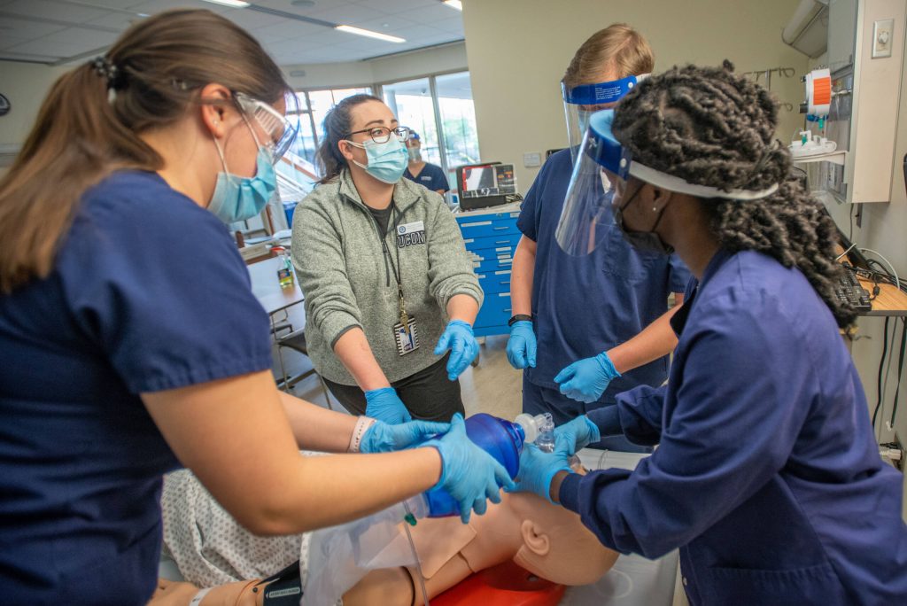 An instructor guiding three students in scrubs on how to save a life by practicing on a dummy