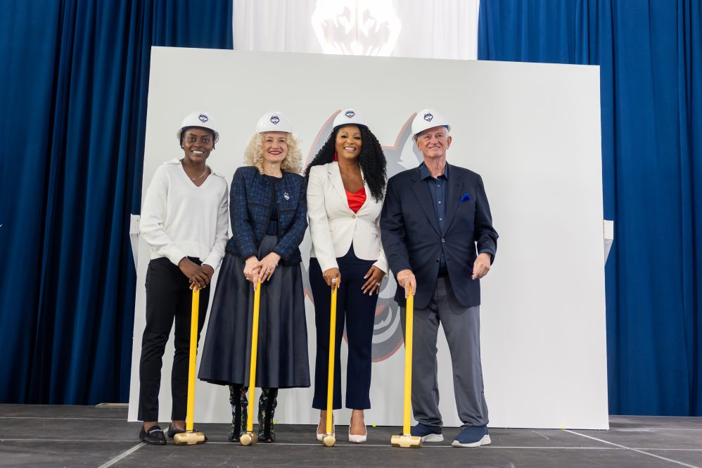 Chioma Okafor, UConn President Radenka Maric, Trisha Bailey, and Denis Nayden posing onstage at the groundbreaking ceremony with UConn branded hard hats.
