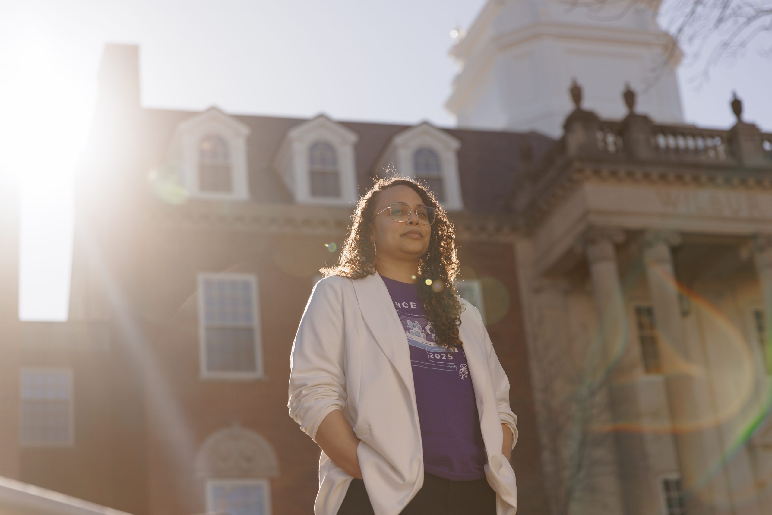 Stephany Santos in a white blazer and purple shirt, standing in front of a campus building, backlit by the sun.
