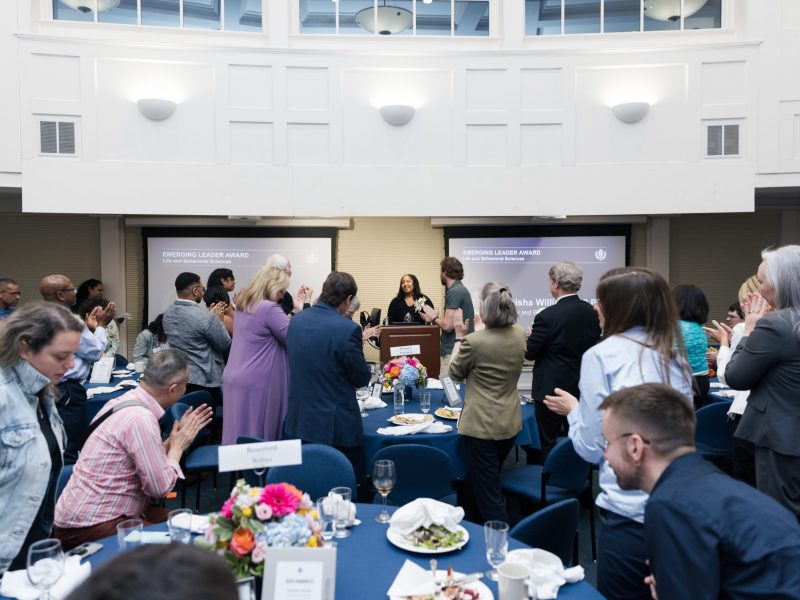Award recipients from the CLAS Alumni Awards gathered in the Alumni Center Great Hall standing and cheering for a speaker behind the podium.