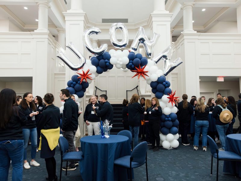 Foundation employees gathered in the Great Hall at the Campaign kickoff event. There is a large balloon arch featuring white and navy balloons, red star-shaped balloons, and silver balloons spelling out "UConn".