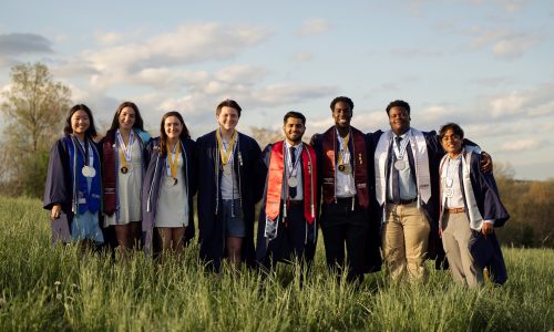 Line of students in graduation regalia smiling with their arms around one another in a grassy field.