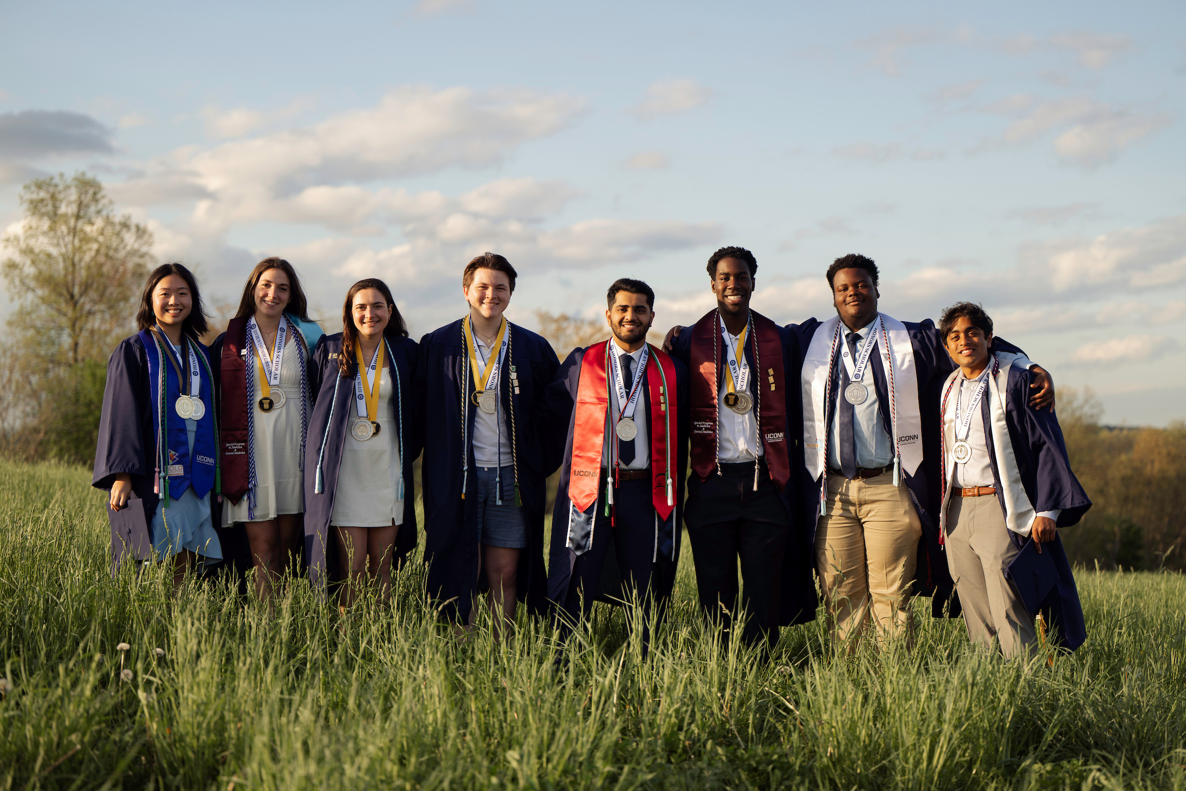 Line of students in graduation regalia smiling with their arms around one another in a grassy field.