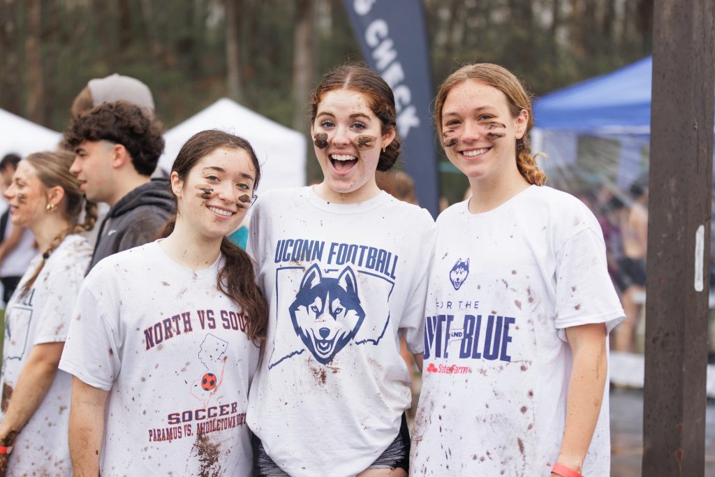 Three girls smiling at the 2025 OOzeball event with white UConn gear and mud on their faces