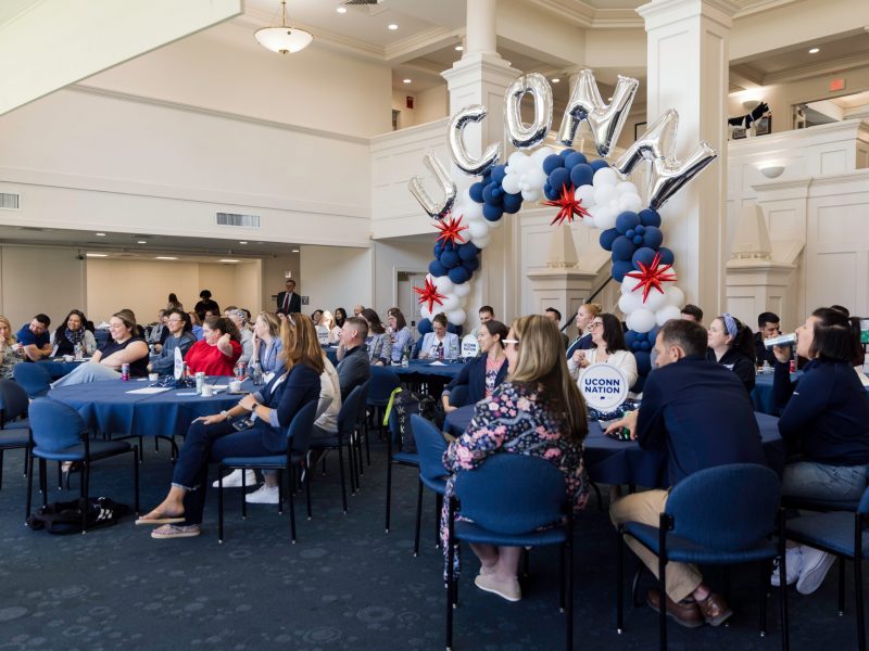 Foundation employees seated at tables in the Great Hall, listening to a panel of students. There is a large balloon arch featuring silver "UConn" balloons over the main staircase.