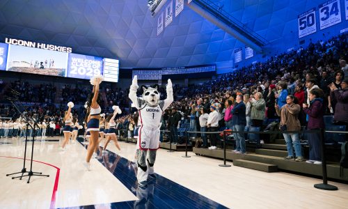 The mascot Jonathan the Husky raising his arms up in Gampel Pavilion in celebration, with the UConn cheer team behind him