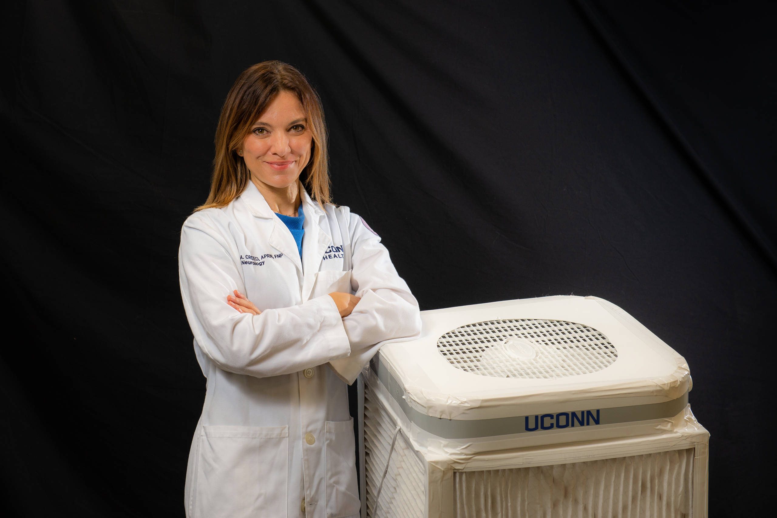 Nurse Practitioner Marina A. Creed crossing her arms and smiling in a UConn Health lab coat, standing next to an air filtration system