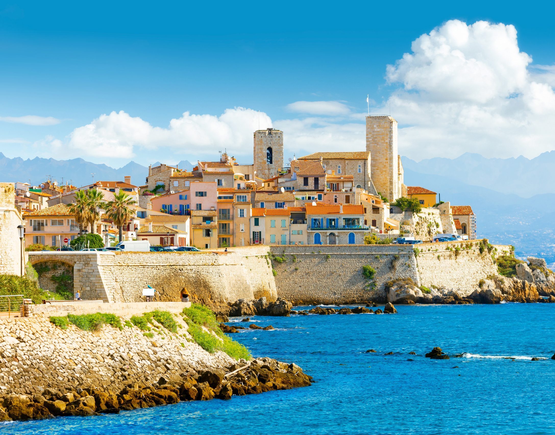 The resort town of Antibes, France on the French Riviera, featuring a rocky coast, brightly-colored orange and pink buildings, a castle structure, and palm trees overlooking turquoise waters.