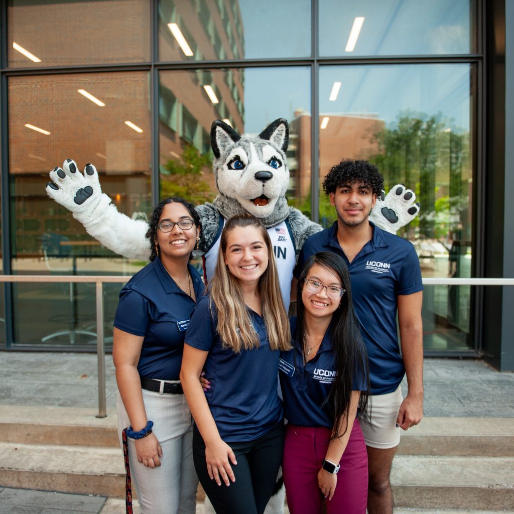 Four smiling students wearing navy blue UConn polo shirts pose together outside a campus building with Jonathan the Husky mascot standing behind them, arms raised at the UConn Pharmacy Alumni Reunion.