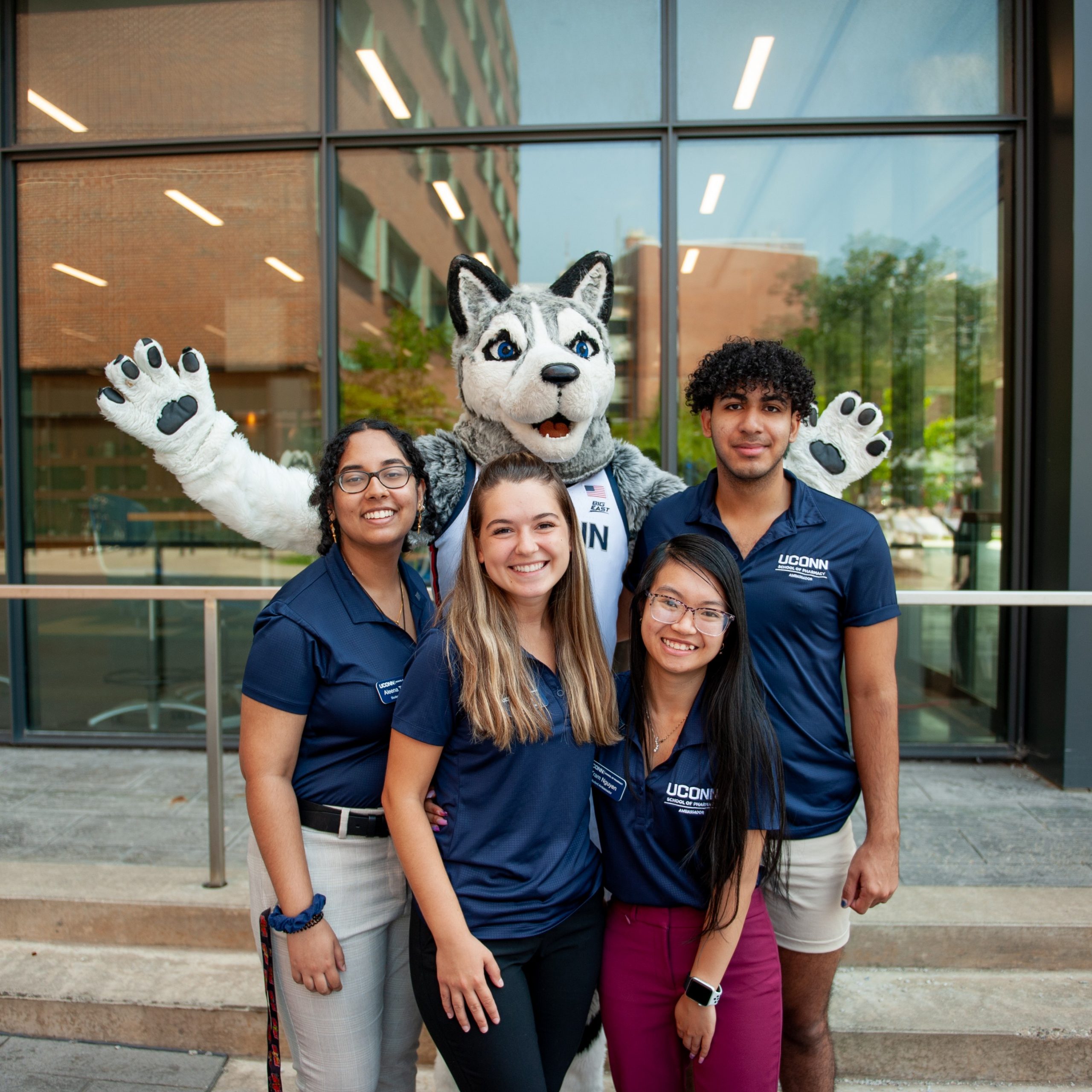 Four smiling students wearing navy blue UConn polo shirts pose together outside a campus building with Jonathan the Husky mascot standing behind them, arms raised at the UConn Pharmacy Alumni Reunion.