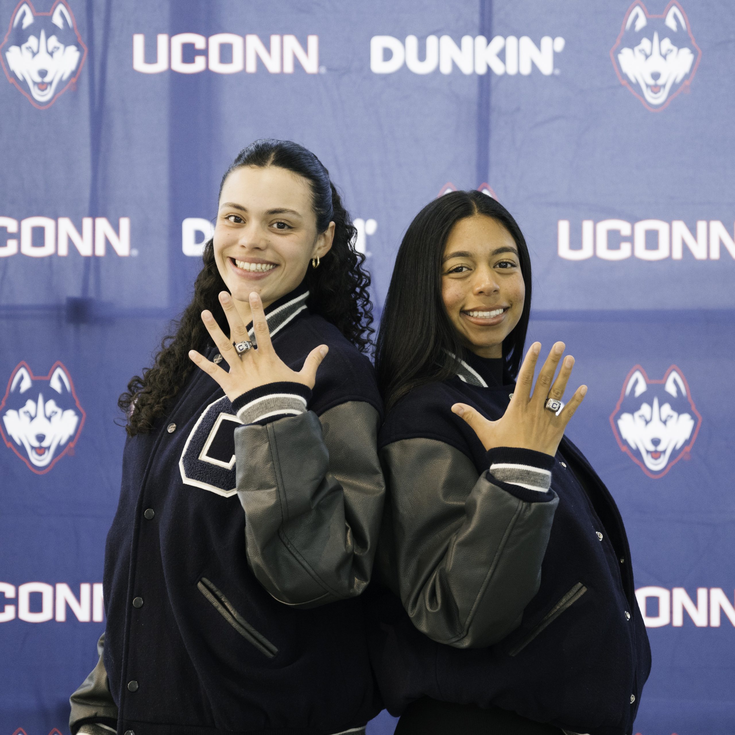 Two UConn athletes wearing UConn varsity jackets smile and proudly displaying rings on their hands. They stand in front of a blue backdrop featuring the UConn Husky logo for the Senior Ring Ceremony.