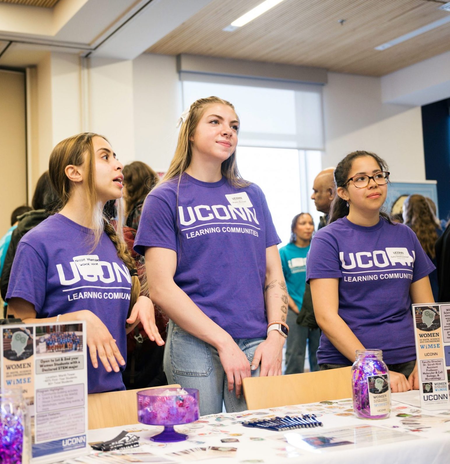 Four students wearing matching purple "UConn Learning Community" t-shirts stand behind a display table at an event or fair. The table is decorated with informational pamphlets, jars, and a banner promoting the "Women in STEM" community. Other attendees can be seen in the background of the brightly lit room.