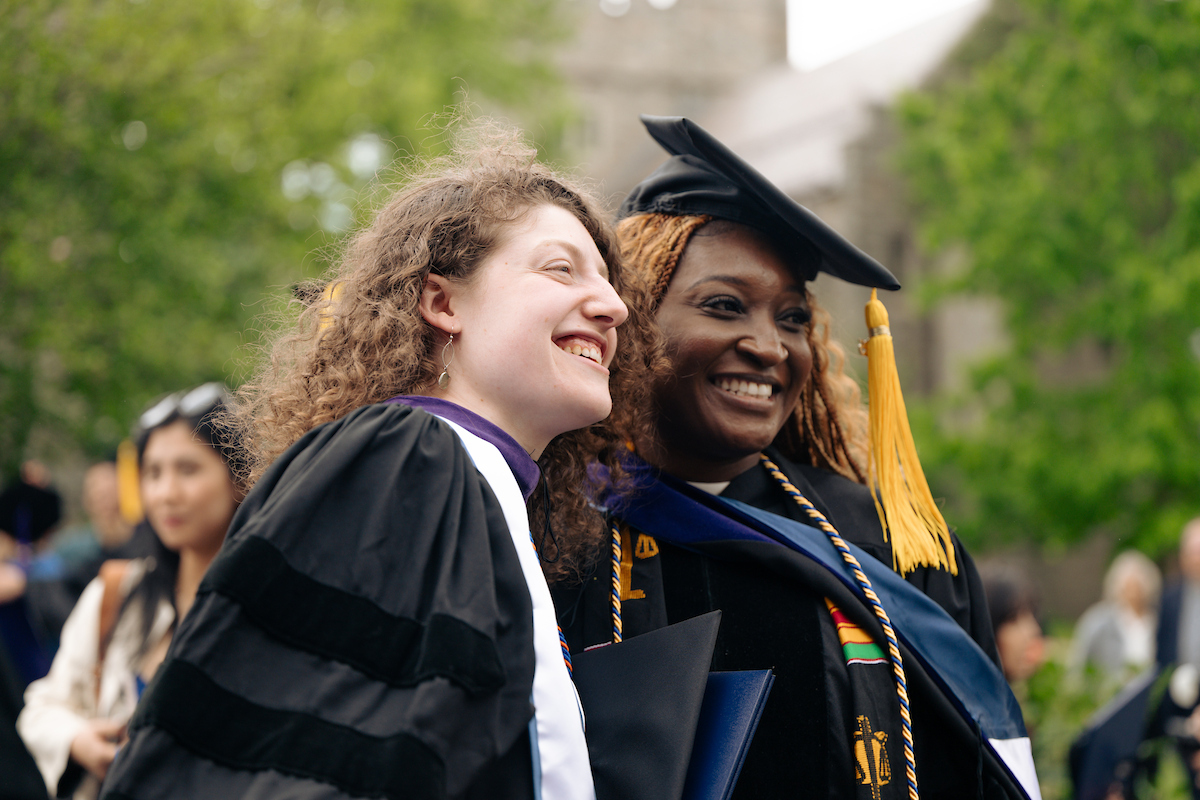Two students in graduation attire smile outdoors.