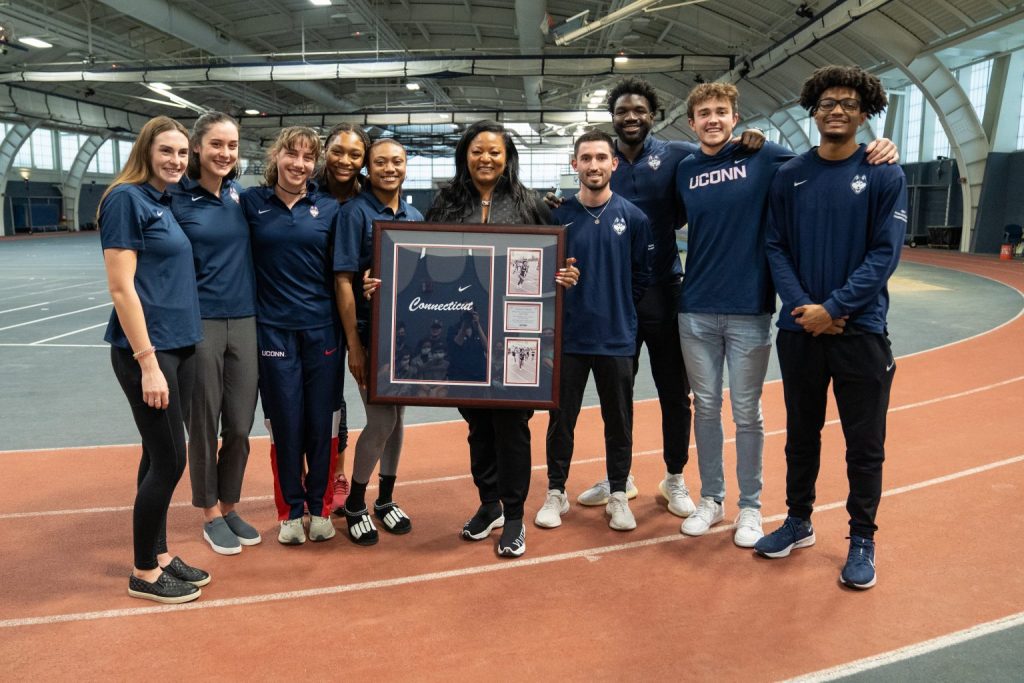 Trisha Bailey pictured with UConn student athletes while she displays her old uniform in the field house