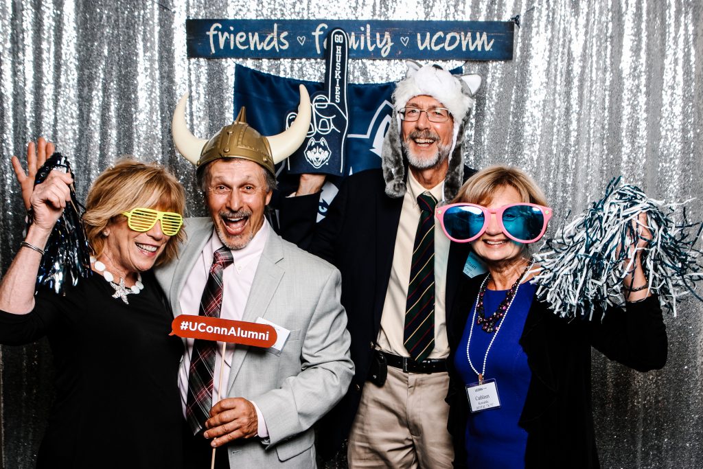 A photobooth photo from the UConn School of Medicine and School of Dental Medicine Reunion featuring people posing with funny props such as oversized sunglasses and a husky hat