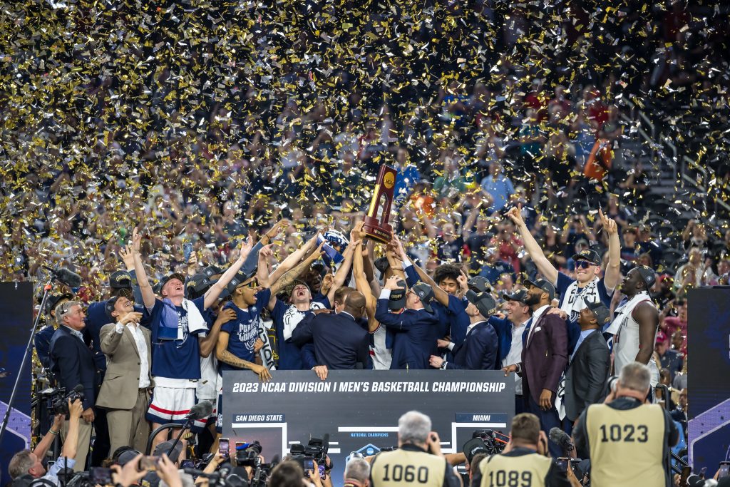 The UConn Men's basketball team celebrating their 2023 championship game with streamers in the air and arms holding up the NCAA trophy