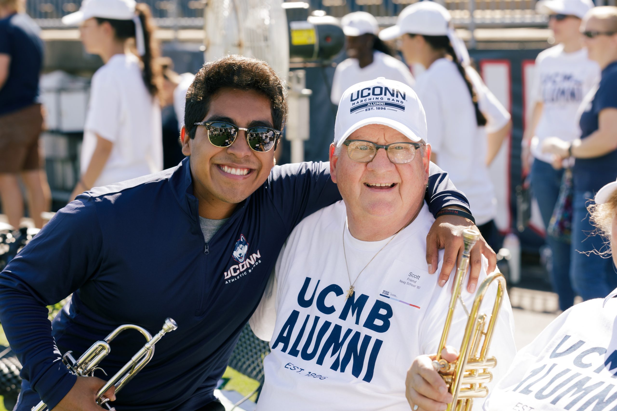 A marching band student and alum posing for a photo while holding their trumpets at the UConn Marching Band Homecoming