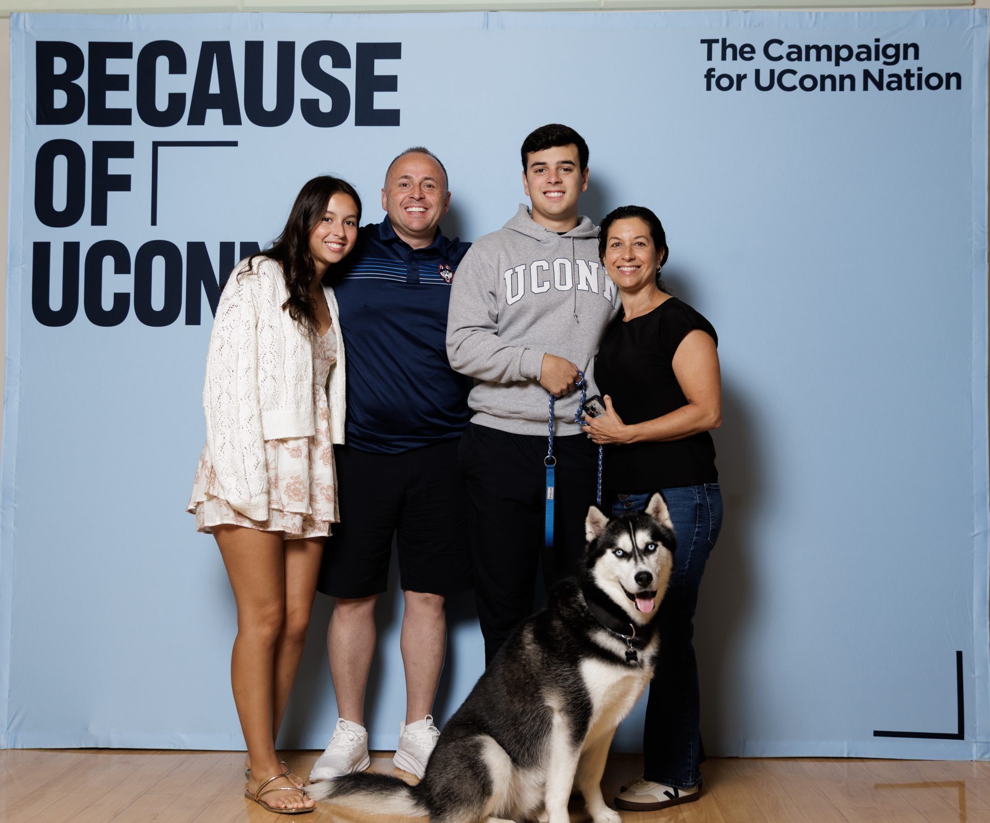A family posing with Jonathan the Husky with a "Because of UConn" backdrop at the Legacy Breakfast