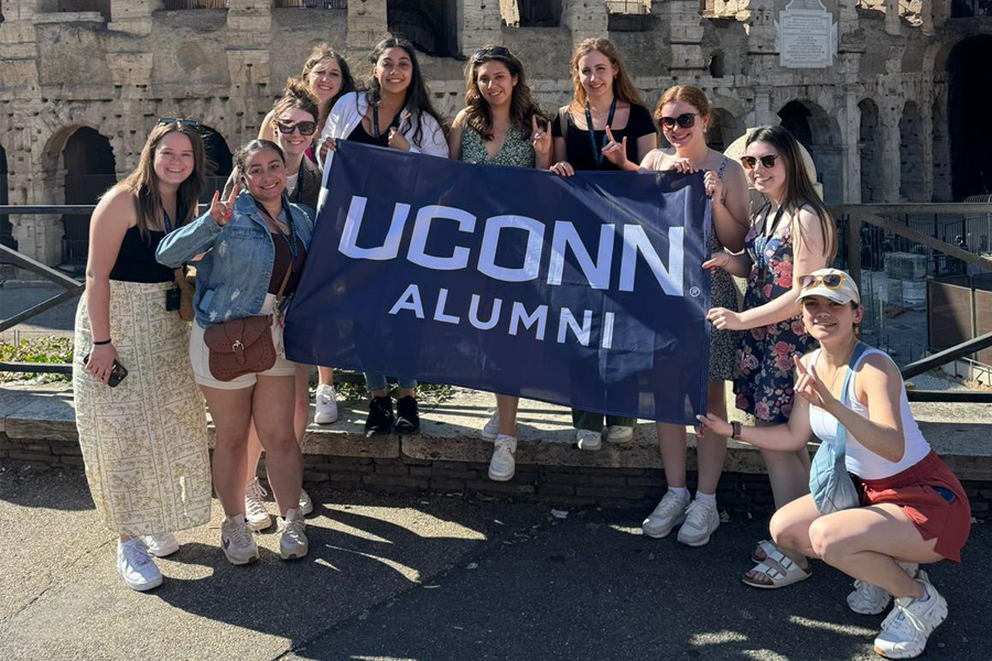 A group of UConn travelers behind a colosseum holding up a navy blue "UConn Alumni" flag