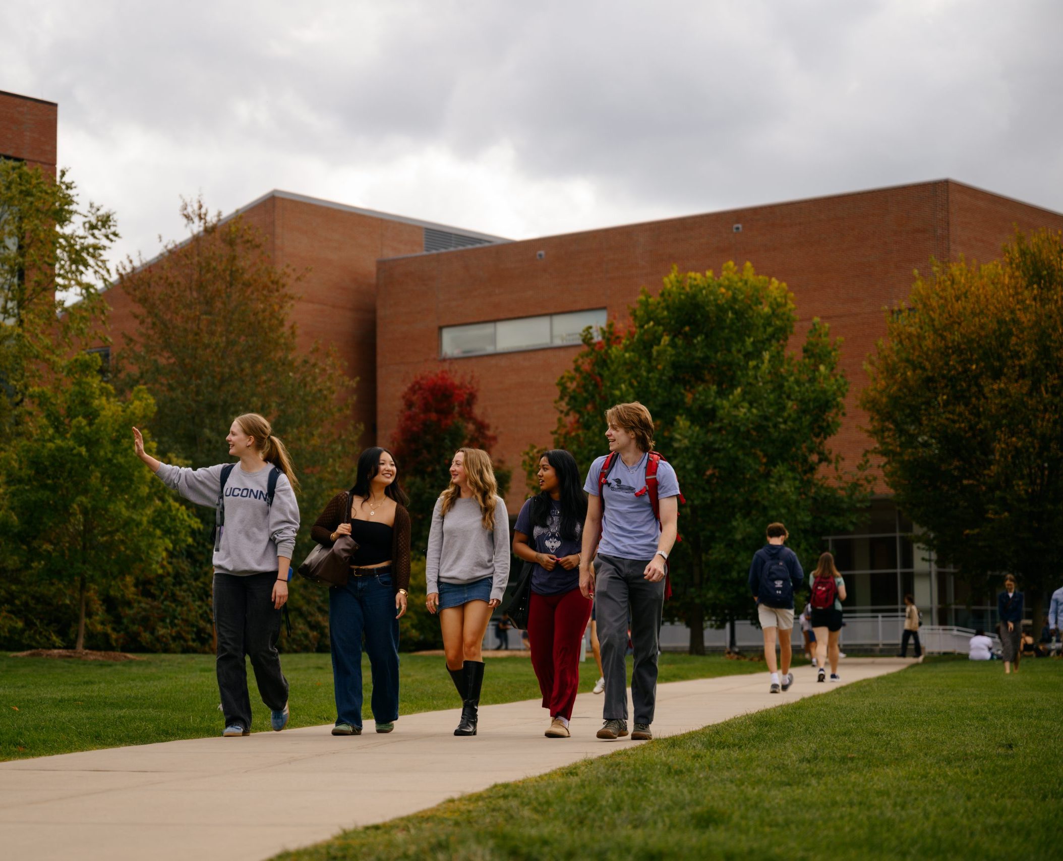 A group of students walk and talk together along the UConn Student Union lawn, with brick academic buildings and trees with early autumn foliage in the background