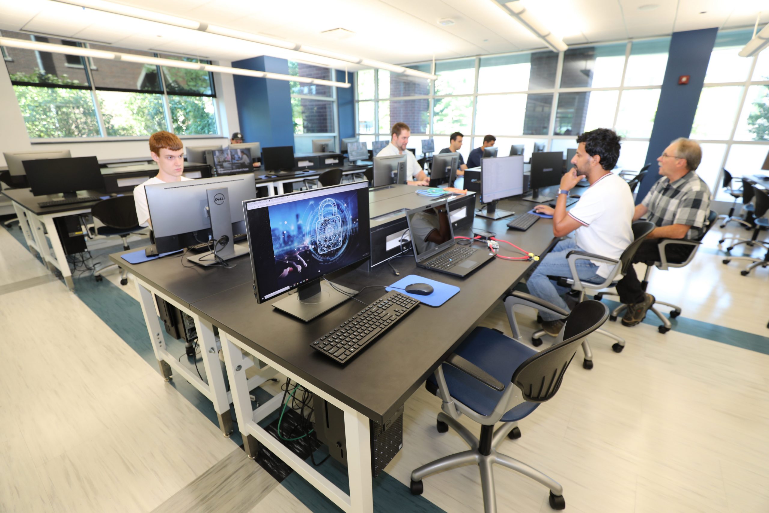 Students work at computer stations in a bright, modern lab with large windows. Multiple monitors display technical graphics as the group collaborates in a spacious classroom setting