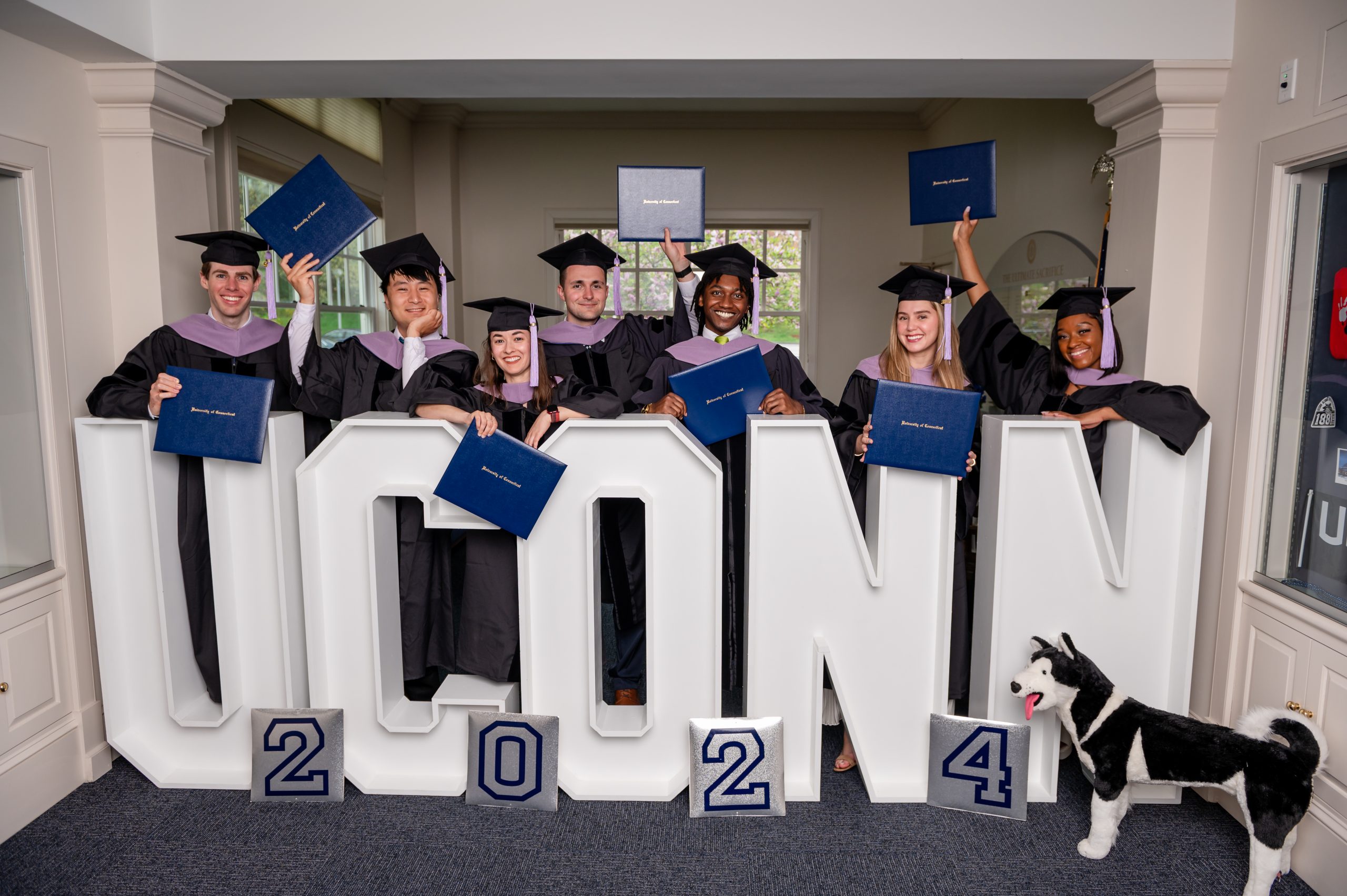 A group of graduates in caps and gowns pose indoors behind large white letters spelling ‘UCONN,’ with smaller numbers reading ‘2024’ in front. They hold up their diplomas in celebration, and a Husky mascot plush stands beside the display