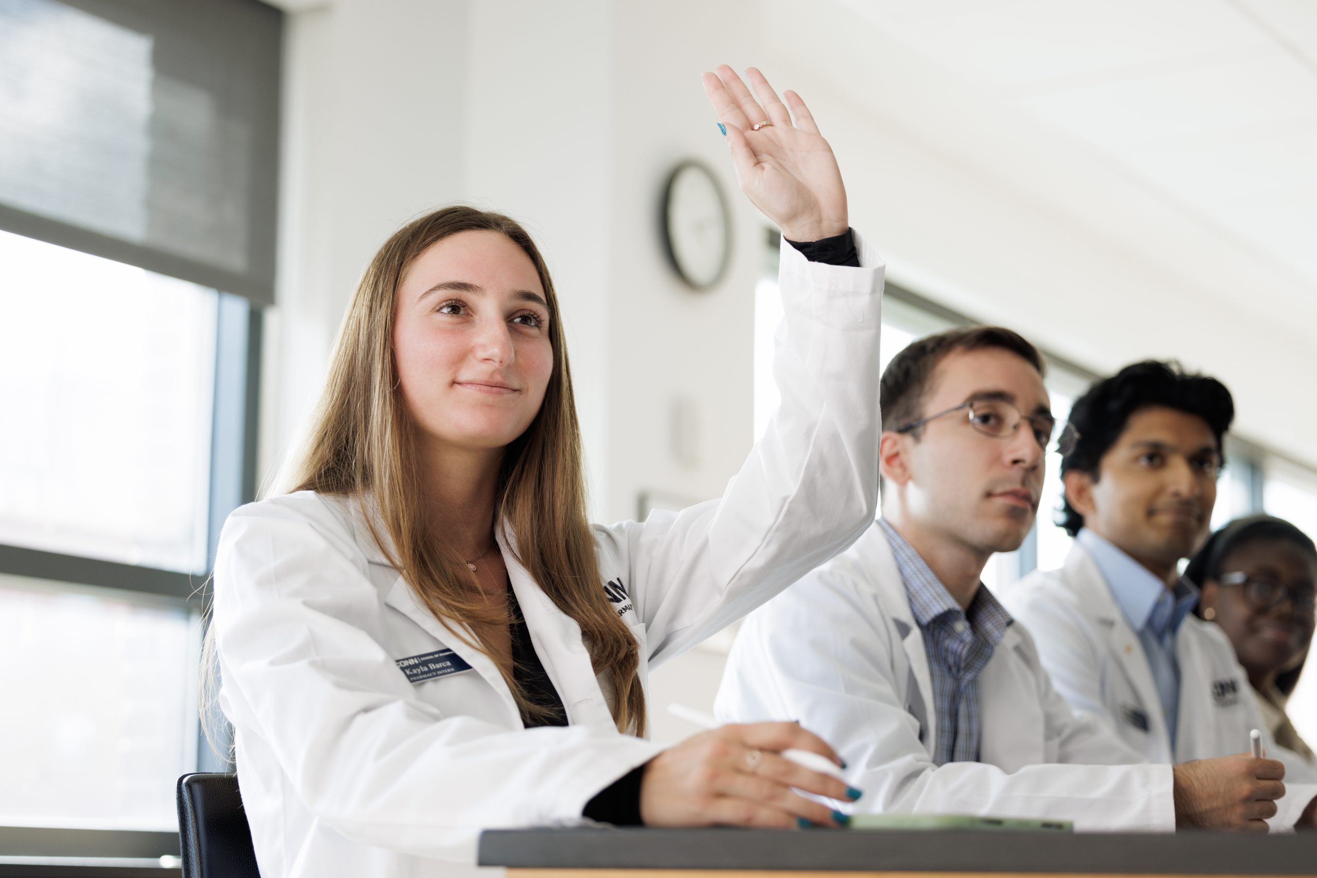 A Pharmacy student in a white lab coat raises a hand during a classroom or lab session, seated among other students in white coats at desks