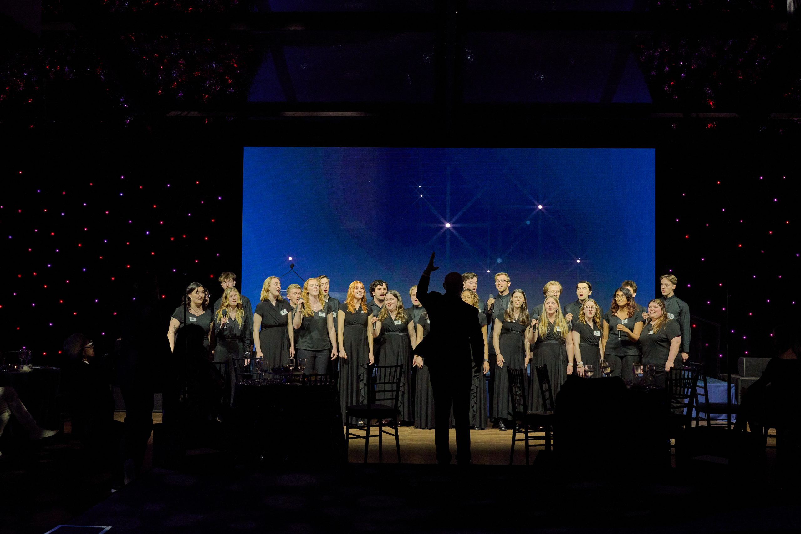 A choir dressed in black performs onstage in front of a large blue backdrop with star-like lights, while a conductor stands in silhouette directing them