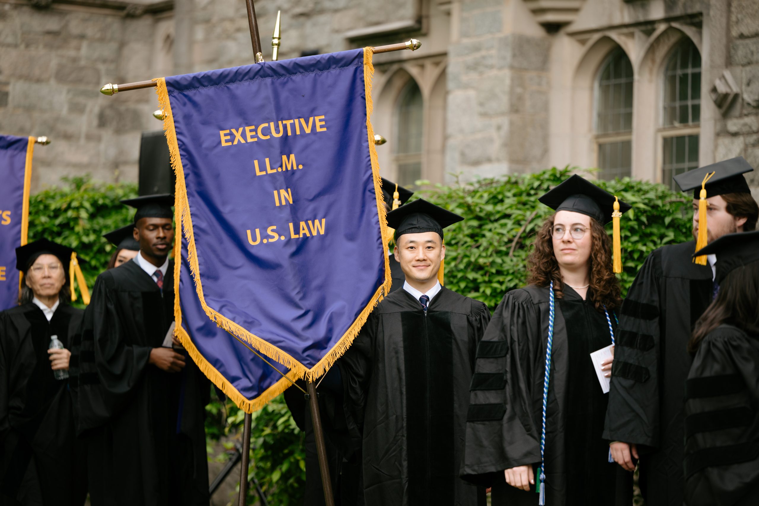 UConn Law graduates in black academic gowns and caps stand outdoors beside a large purple and gold banner that reads ‘EXECUTIVE LL.M. IN U.S. LAW,’