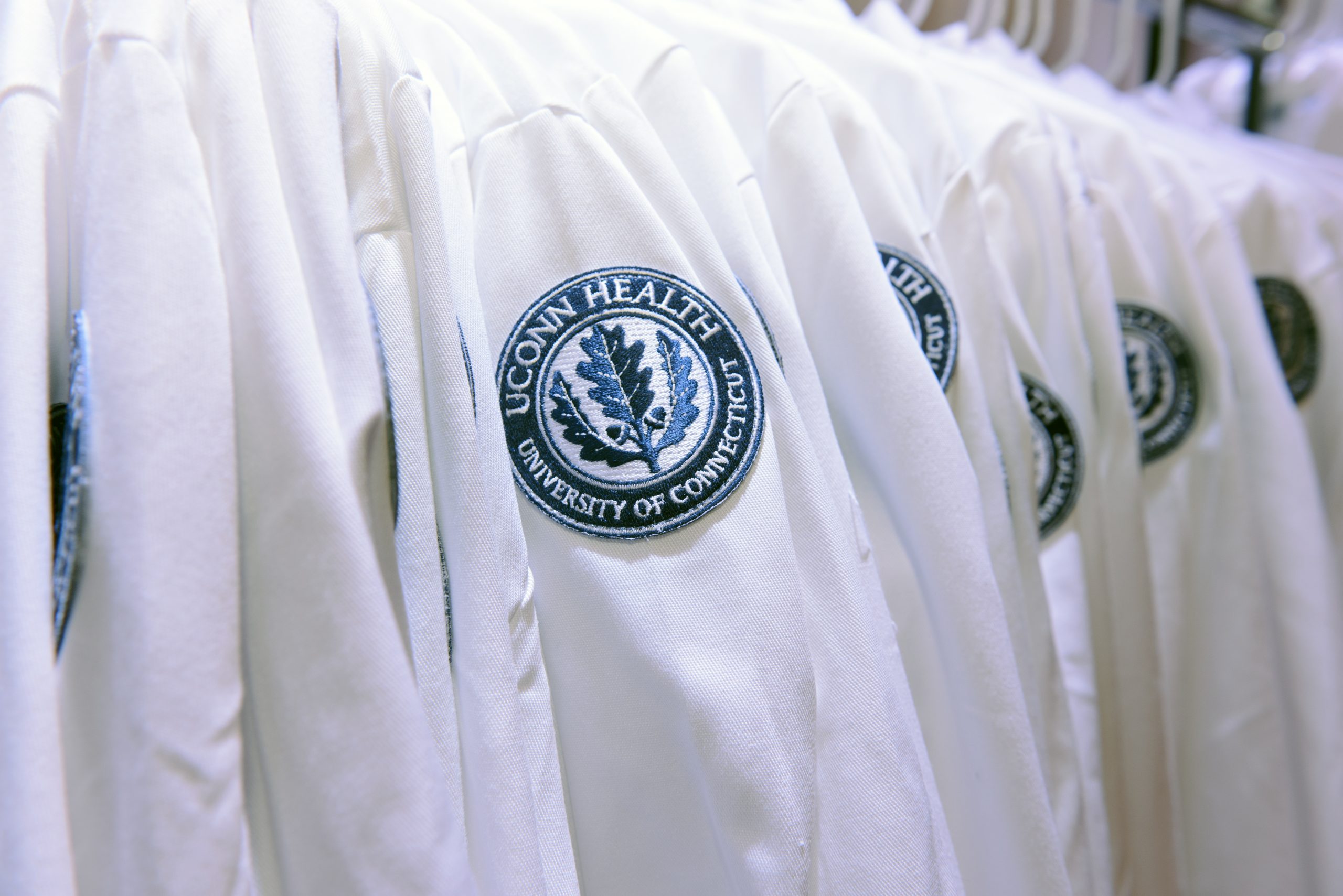 A row of white lab coats hangs on a rack, each embroidered with a UConn Health, University of Connecticut emblem