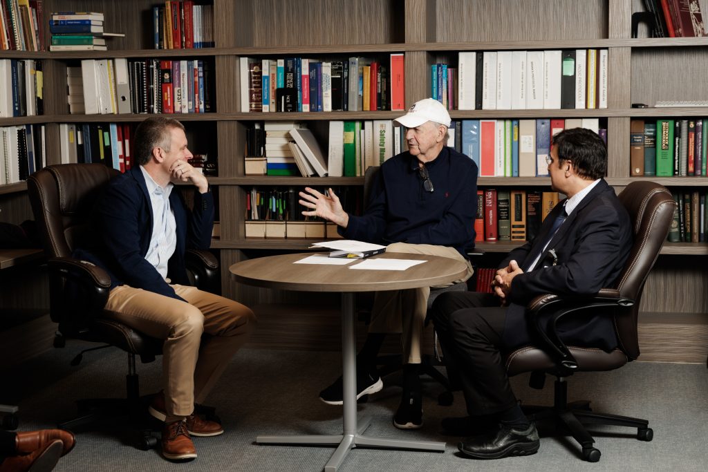 Bulsara, Piasio, and Jim Calhoun seated together and in conversation at a round table with bookshelves in the background.