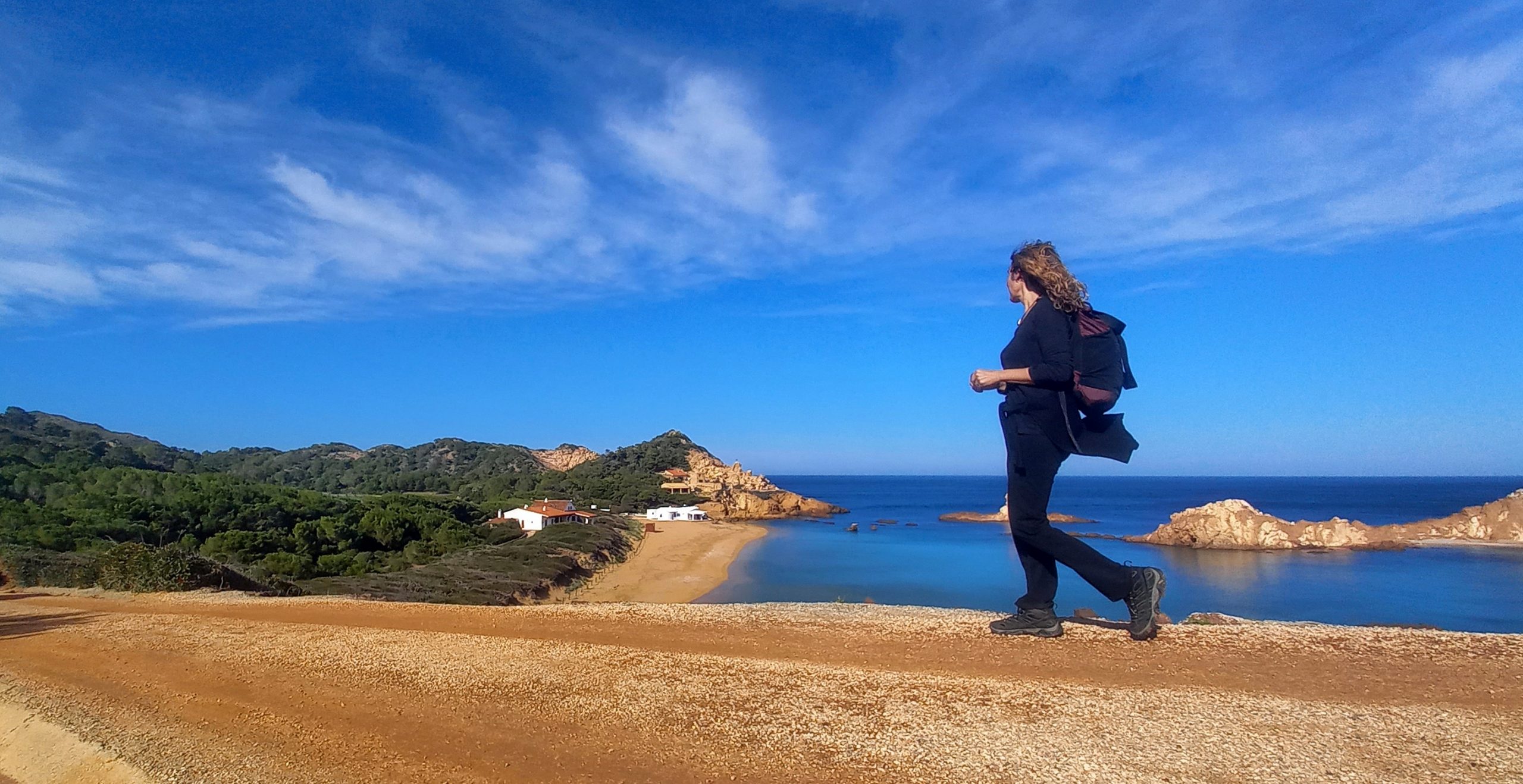 A woman walking on a dirt path overlooking a small bay with bright blue water in Menorca