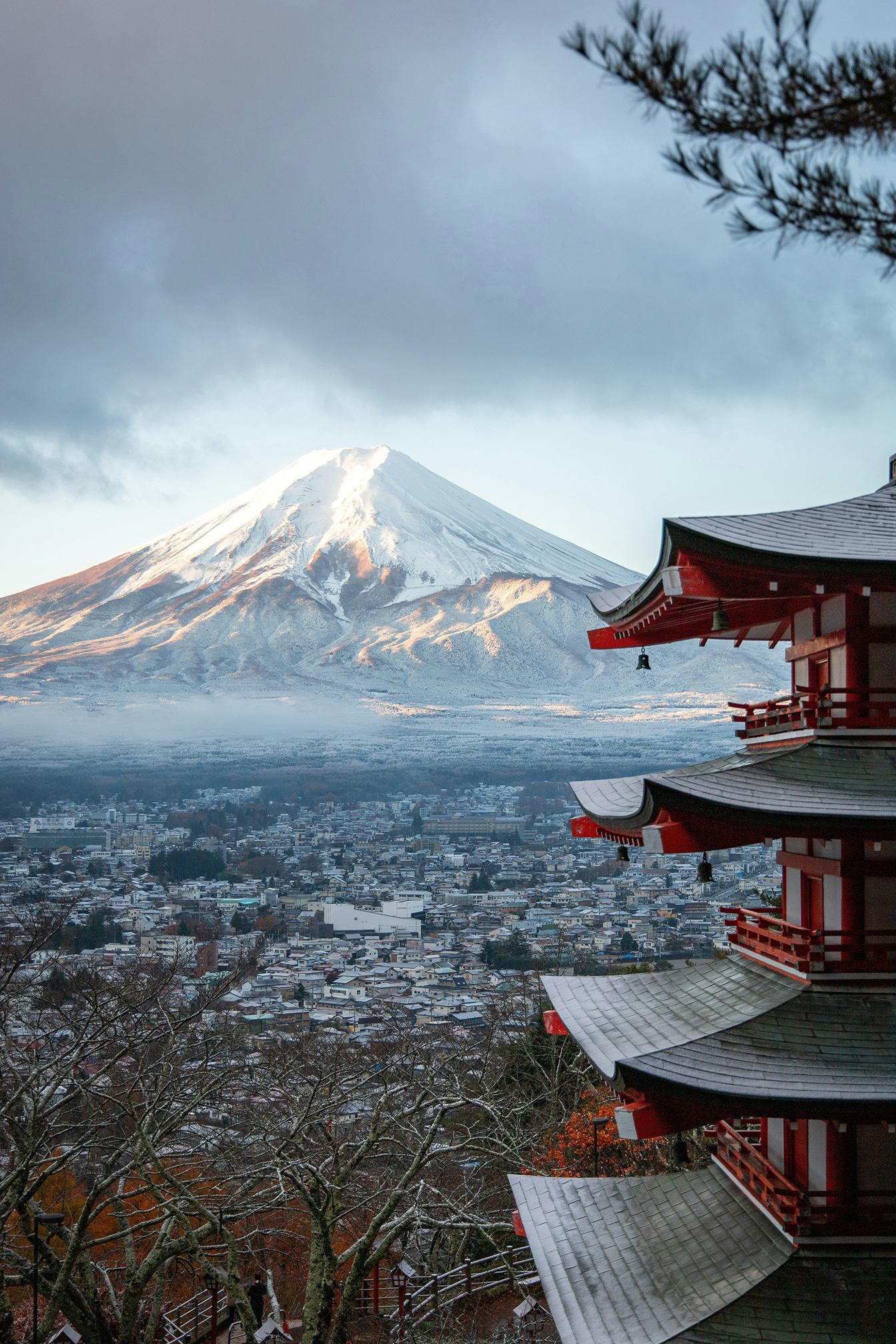 Snow-capped Mount Fuji rises above a small town, with a red multi-tiered pagoda in the foreground and soft morning light illuminating the mountain and clouds.