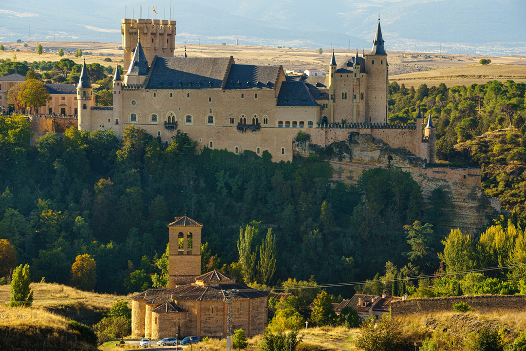 Historic stone castle perched on a wooded hilltop overlooking a small church and rural landscape, with towers, spires, and fortified walls set against distant mountains under clear daylight.