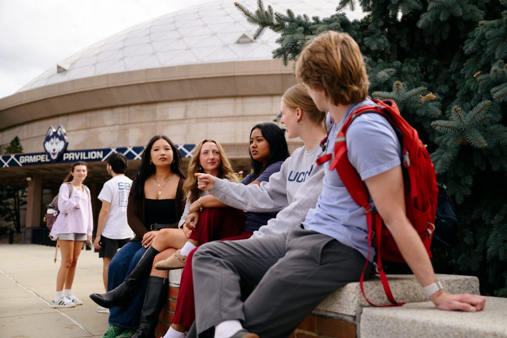 Students sitting on a stone wall and chatting, with Gampel Pavilion seen in the background