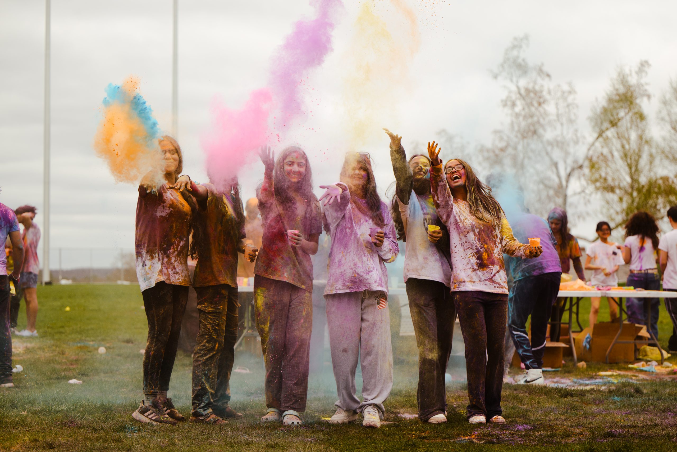 Participants at a Holi celebration toss colorful powder into the air while standing together on a grassy field.