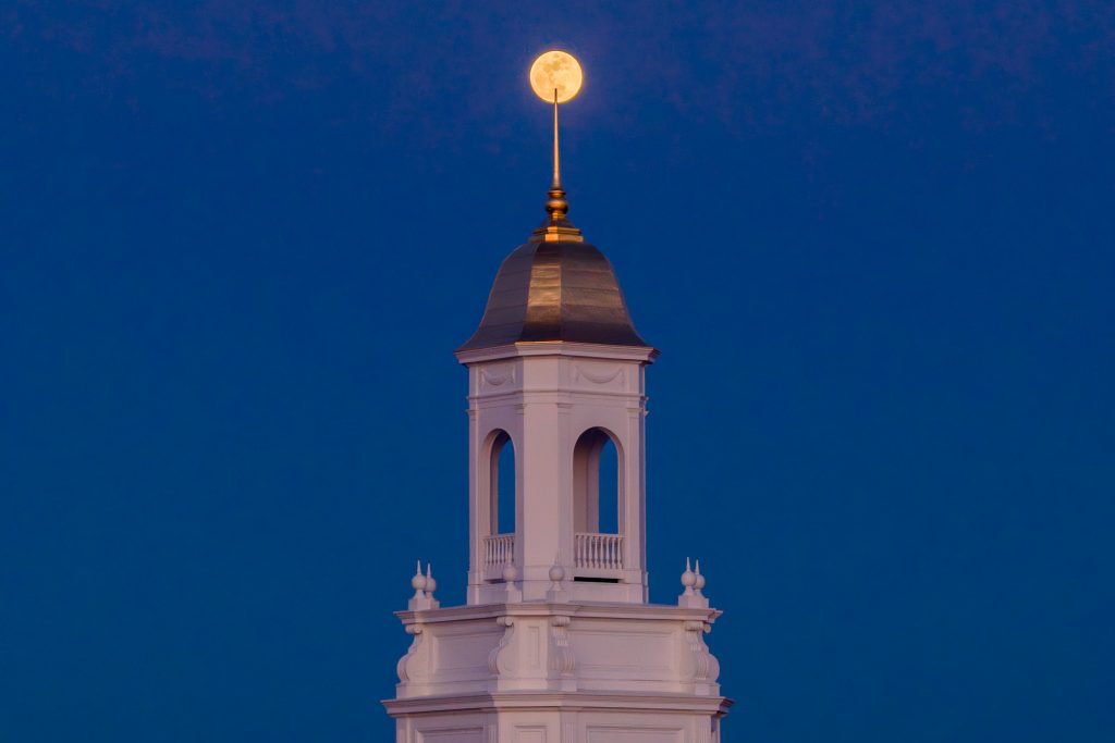 The top of the UConn campus cupola stands against a deep blue evening sky, with a full moon appearing perfectly aligned above the spire.